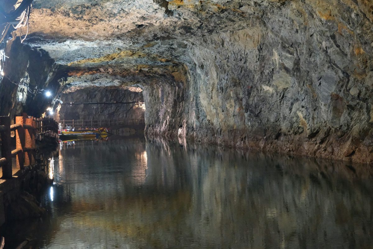 A flooded underground mine gallery lit along the rock wall, with boats moored at a timber walkway