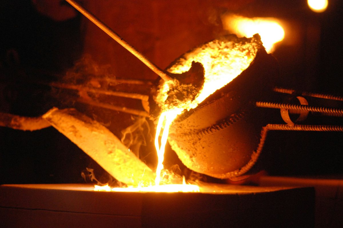 Molten metal pouring from a tilted crucible into a mould, lit orange in a dark foundry